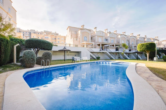 Fotografía principal de un amplio adosado moderno de varias plantas en la urbanización Golf de Alicante. Se muestra su fachada blanca, terrazas en diferentes niveles, zona de barbacoa y acceso a la piscina comunitaria, con jardín y palmeras